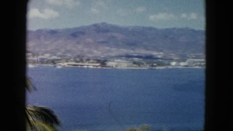 1962 Acapulco home-movie view across open bay water toward a coastal strip and mountains, with a palm edge at the lower left.