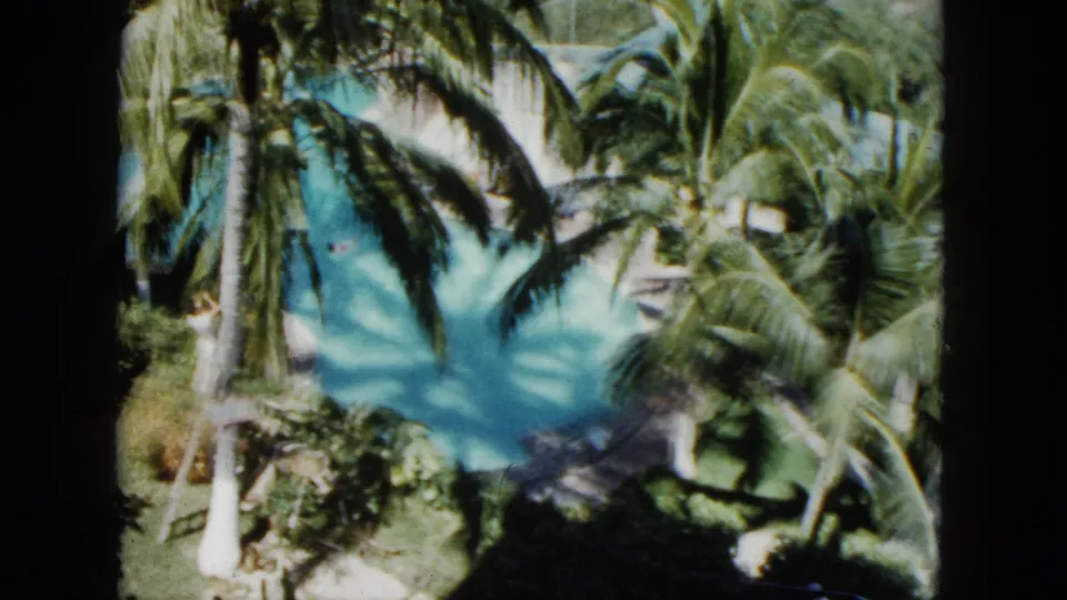 1962 Acapulco home-movie frame looking down at a turquoise pool surrounded by palms and sunlit garden planting.