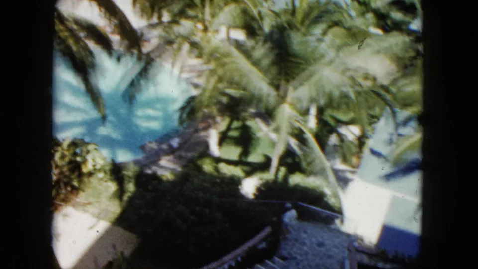 1962 Acapulco 8mm frame looking over palms, pool water, a white wall, and a shaded curved walkway.