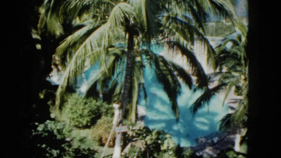 1962 Acapulco 8mm still with large palm fronds, dense greenery, and aqua swimming pool water under soft, grainy exposure.