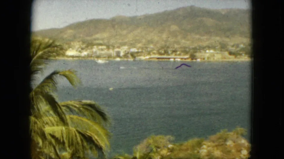 1962 Acapulco home-movie view of a bay, distant hillside town, palm fronds, and hazy mountains beyond the water.