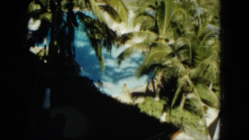 1962 Acapulco home-movie frame of a tropical pool area seen through palms, with small swimmers or visitors near the water.