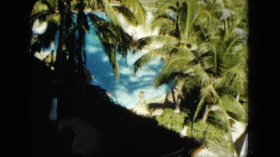 High-angle 1962 Acapulco resort pool view with palms, bright blue water, shaded foreground, and small people visible near the pool.