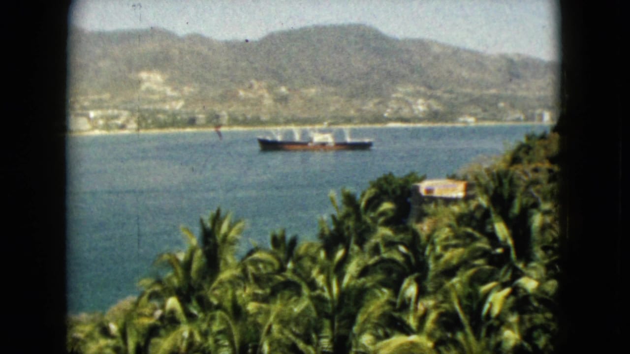 1962 Acapulco harbor view through a dark window frame, with palms in the foreground, a large ship on blue water, and mountains behind the town.
