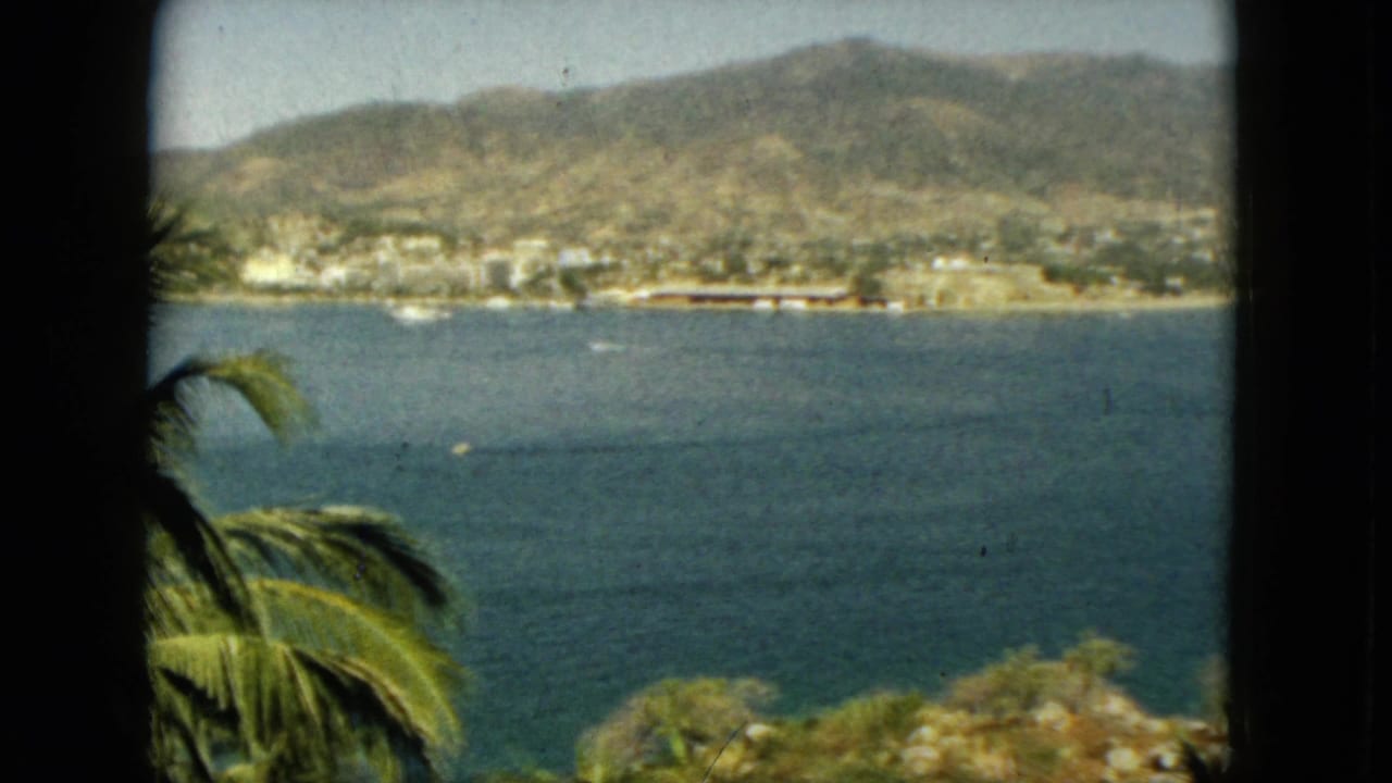 1962 Acapulco bay seen from a shaded opening, with palm fronds below, open water, small boats, waterfront structures, and mountains in the distance.