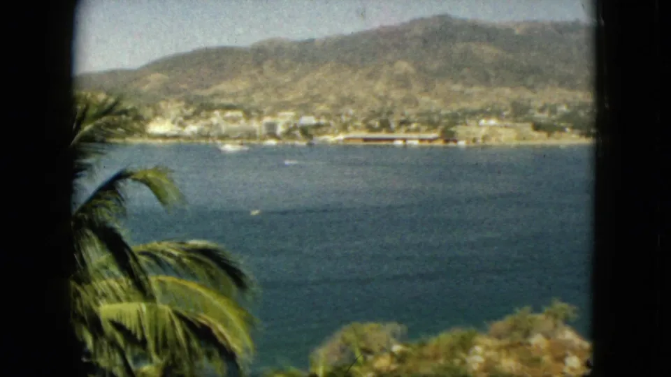 1962 Acapulco bay view with tropical foliage in the foreground, small boats on the water, a waterfront town, and dry mountain slopes behind it.