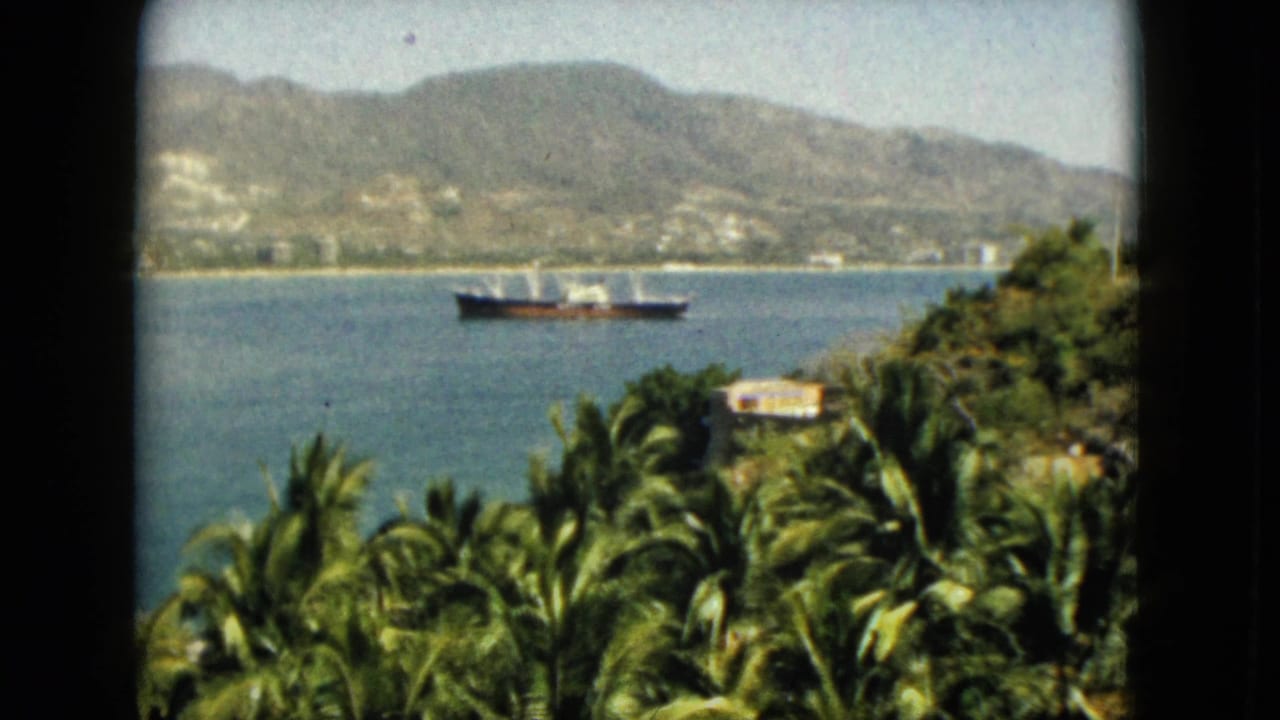 1962 Acapulco 8mm frame showing a cargo ship on the bay, framed by palm trees and a distant mountain shoreline.