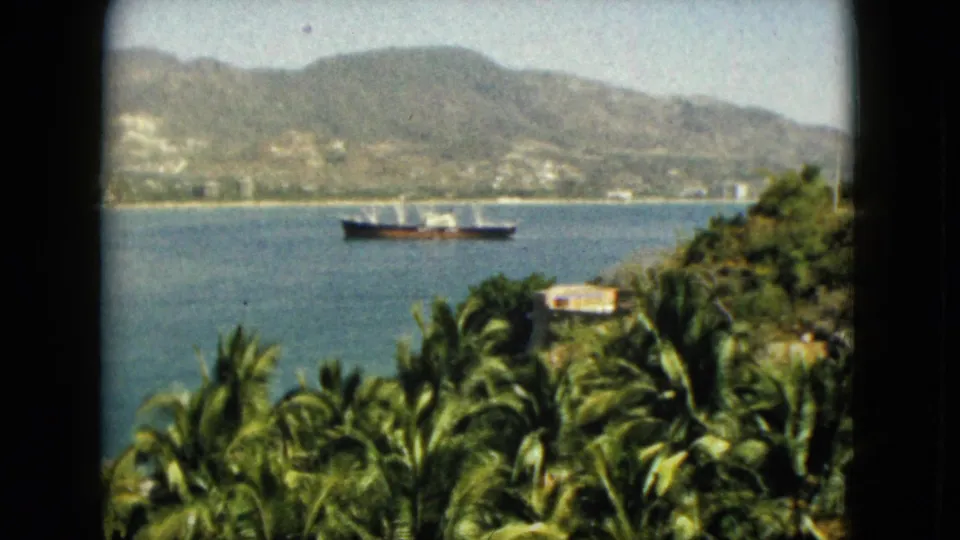 1962 Acapulco 8mm frame showing a cargo ship on the bay, framed by palm trees and a distant mountain shoreline.