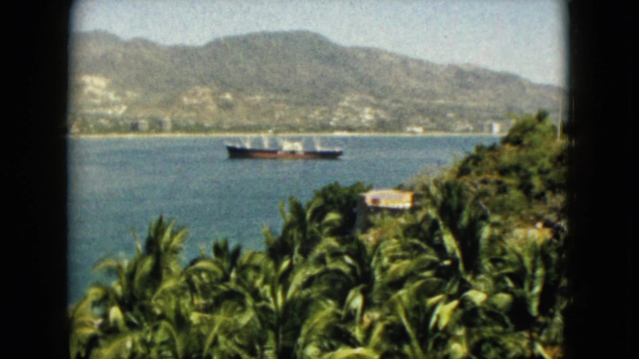 1962 Acapulco home-movie view of a ship moving across calm water with palms, hillside greenery, and mountains in the background.