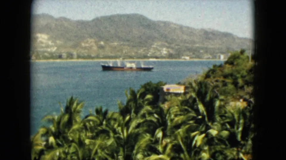 1962 Acapulco home-movie view of a ship moving across calm water with palms, hillside greenery, and mountains in the background.