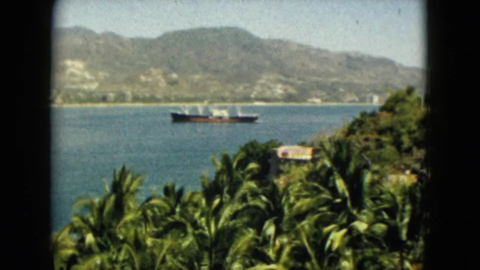 Soft 8mm frame from Acapulco in 1962 with a cargo ship offshore, palm trees below, and a hazy mountain-backed coastline.