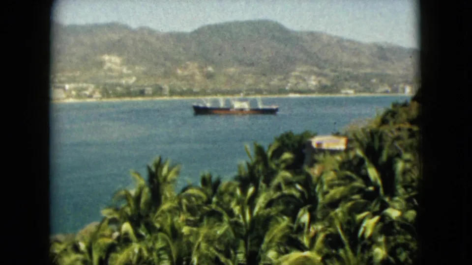 1962 Acapulco bay view showing a distant ship, tropical palms, a small hillside structure, and mountains across the water.