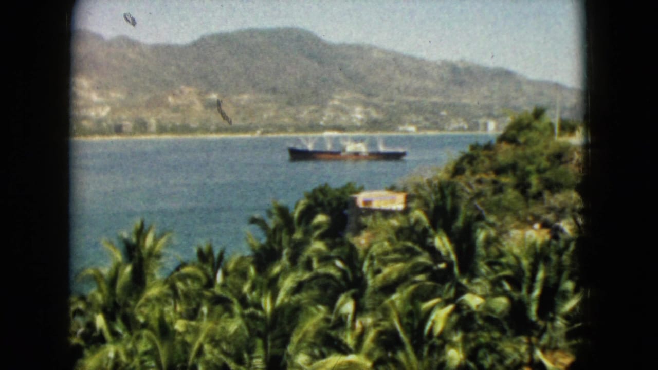 1962 Acapulco home-movie frame showing palms in the foreground, a ship on the bay, distant shoreline buildings, and mountains.