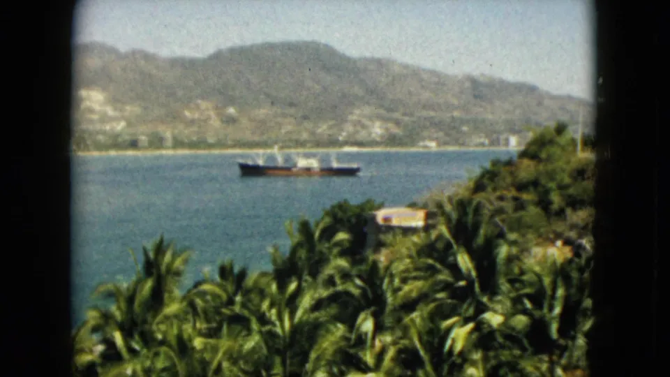 1962 Acapulco frame from an elevated viewpoint, with palms, a small building, bay water, a ship, and hazy hills in the distance.