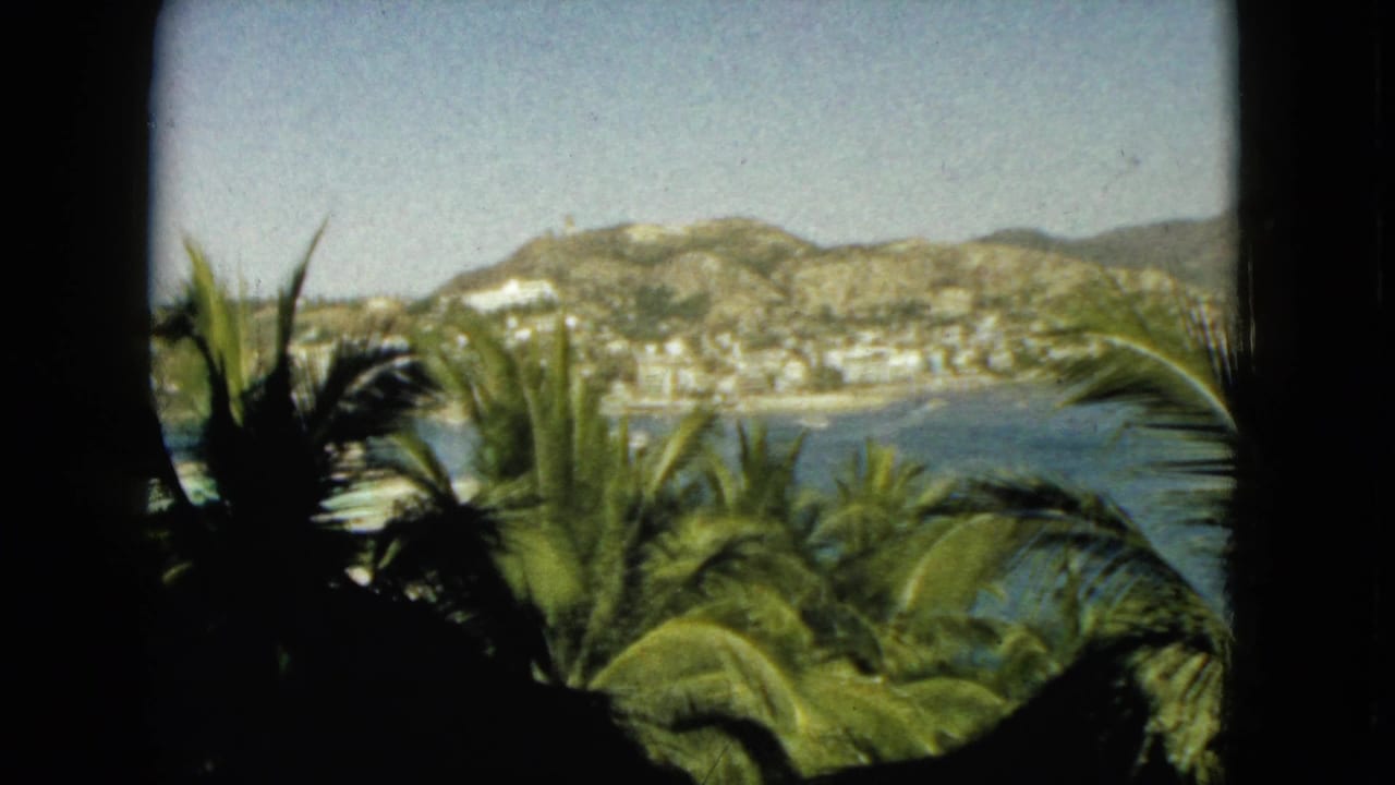 1962 Acapulco home-movie view through a dark window frame, showing palm fronds in the foreground, blue bay water, hillside buildings, and bright sky.
