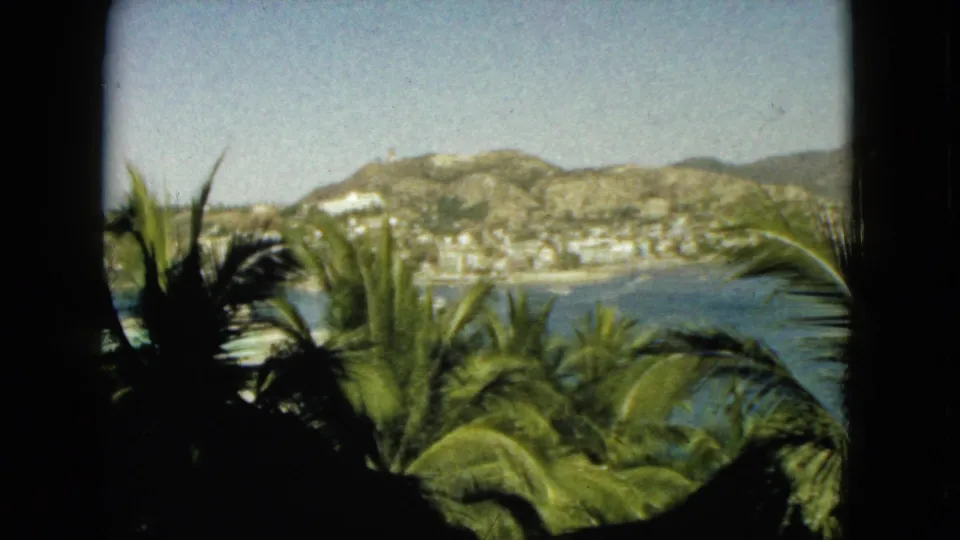 1962 Acapulco home-movie view through a dark window frame, showing palm fronds in the foreground, blue bay water, hillside buildings, and bright sky.