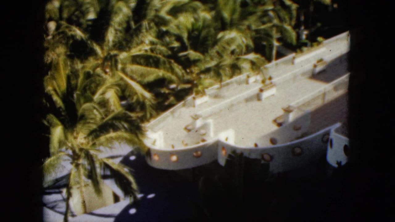 1962 Acapulco frame showing a white resort structure with round openings beside dense palm foliage.