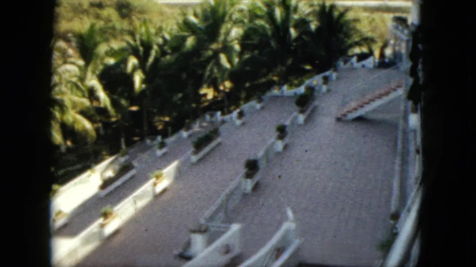 1962 Acapulco home-movie frame showing a white resort terrace with palms, planters, tiled paving, and curved nautical-style railings.