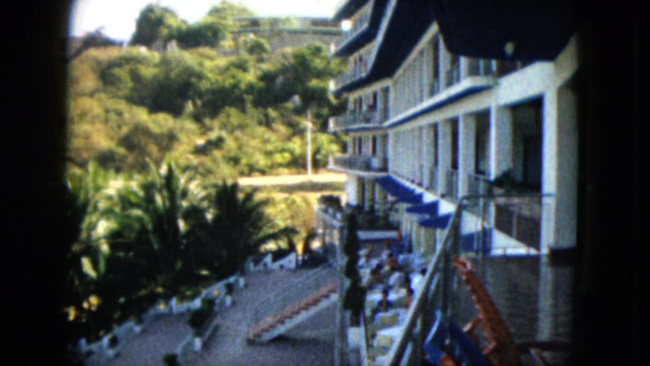 1962 Acapulco home-movie frame showing a white hotel with balconies above a sunny terrace, palms, hillside, and distant guests.