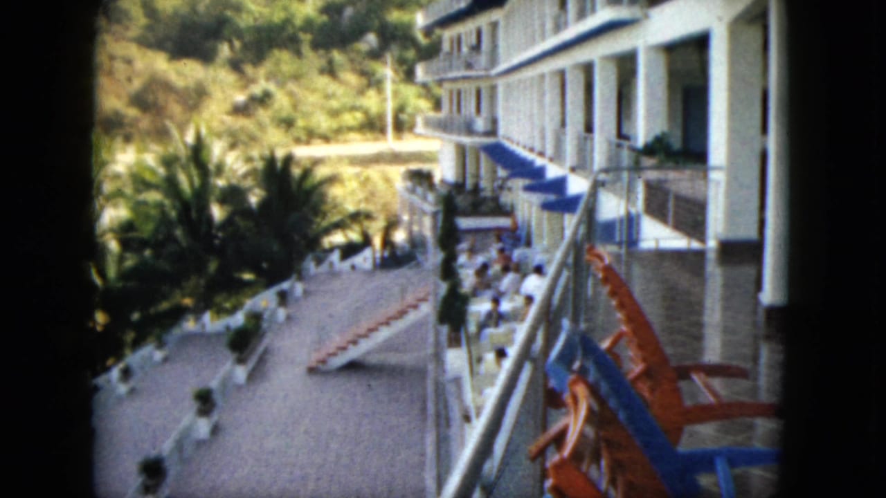 White hotel facade and terrace with blue awnings, palms, chairs, and guests.