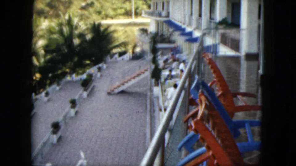 1962 Acapulco frame looking down past balcony railings to a tiled hotel terrace, palms, blue awnings, lounge chairs, and distant guests.