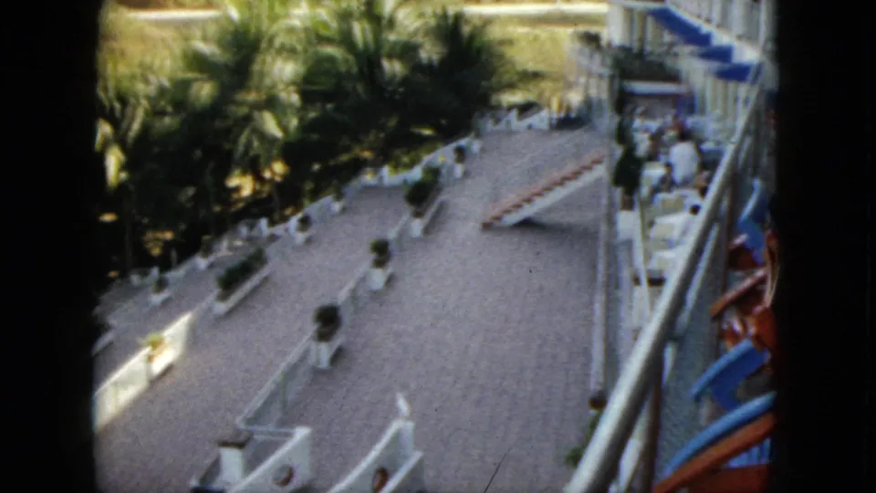 1962 Acapulco home-movie view of a hotel terrace lined with palms and white railings, with lounge chairs and distant guests near the building.
