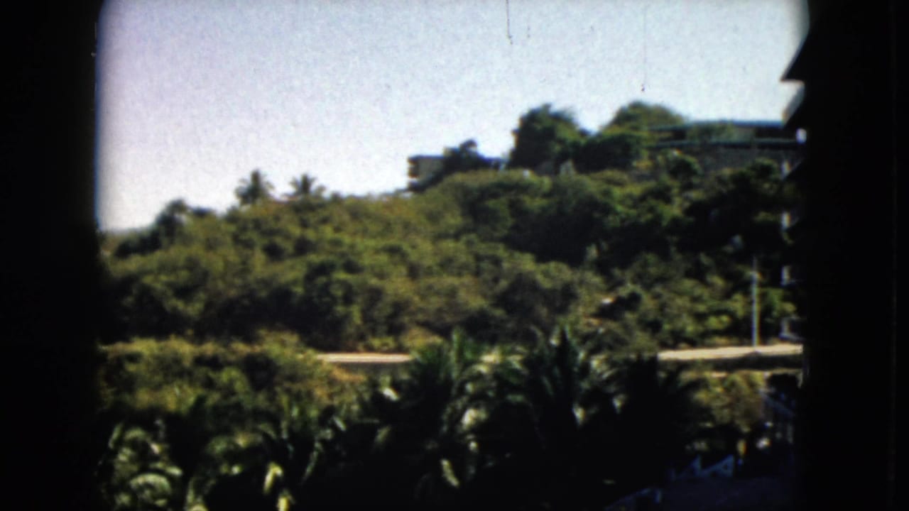 1962 Acapulco view of dense hillside greenery, a pale road, bright sky, and distant buildings within a shadowed 8mm frame.