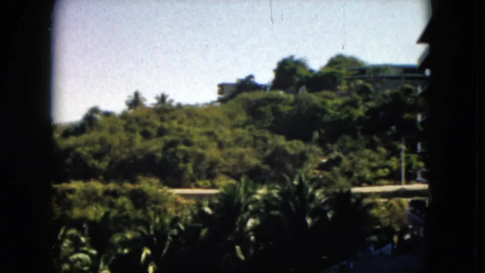 1962 Acapulco view of dense hillside greenery, a pale road, bright sky, and distant buildings within a shadowed 8mm frame.