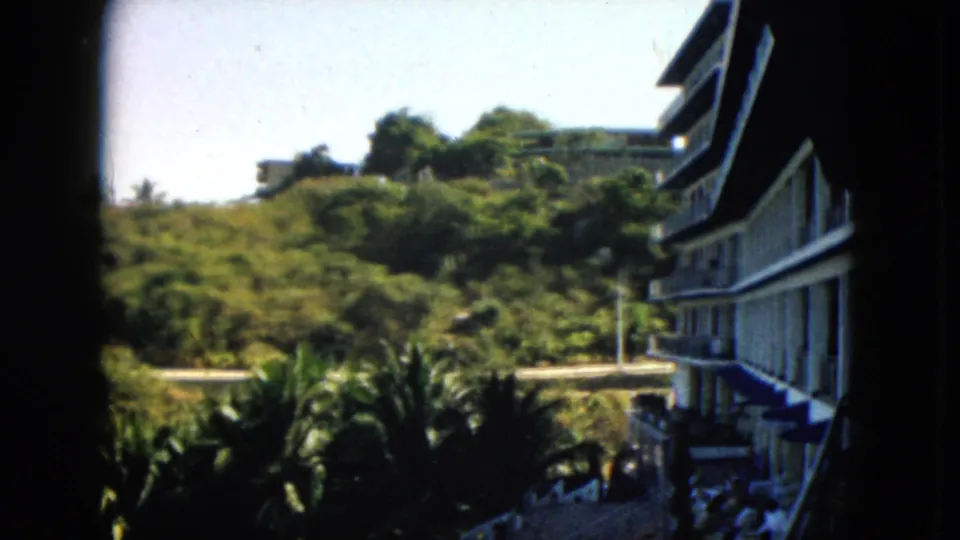 1962 Acapulco 8mm frame with a resort-like balcony building, palms, hillside vegetation, and a curving road.
