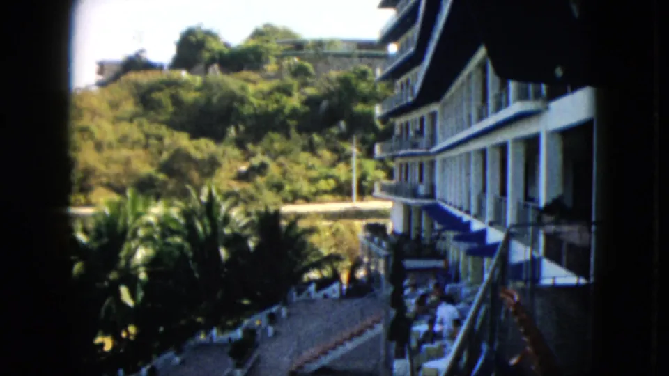1962 Acapulco frame showing a multi-story balcony building, palms, hillside road, and small distant people near the lower right.