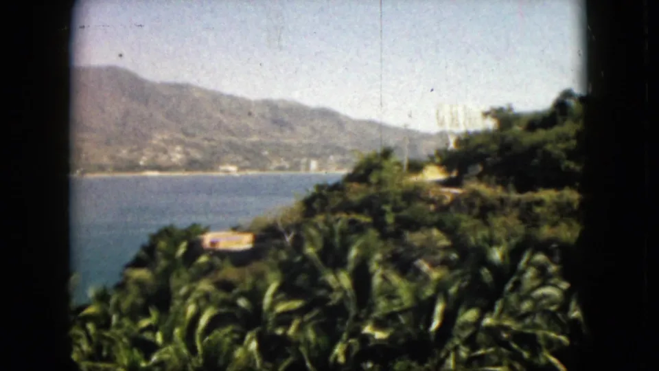 1962 Acapulco coastal view with bay water, distant hillside buildings, palms, and dense greenery at the right edge.