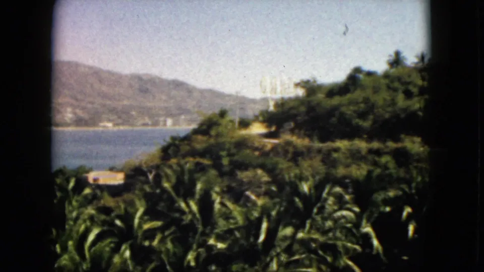 1962 Acapulco home-movie frame with palms below, bay and mountains behind, and a bright hillside feature among trees.