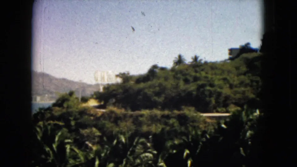 1962 Acapulco coastal hillside view as the pan settles on dense greenery, with bay water and mountains partly visible at left.