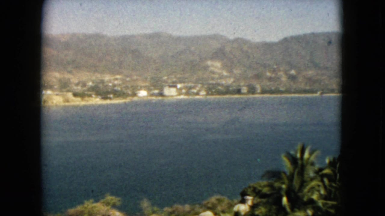 1962 Acapulco home-movie frame showing blue bay water, a distant shoreline, palms, and hazy mountains through dark film edges.