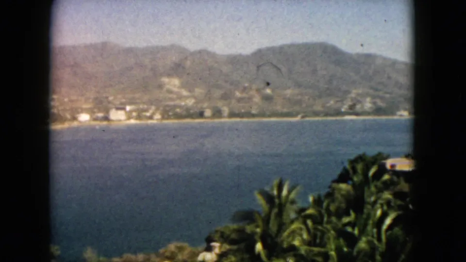 Soft 8mm frame of Acapulco bay, showing blue water, shoreline development, palm tops, and a hazy mountain ridge.