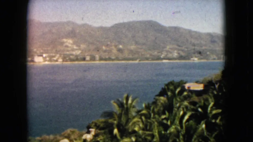 1962 Acapulco bay overlook with foreground palms, dense foliage, blue water, shoreline buildings, and mountains in faded 8mm color.