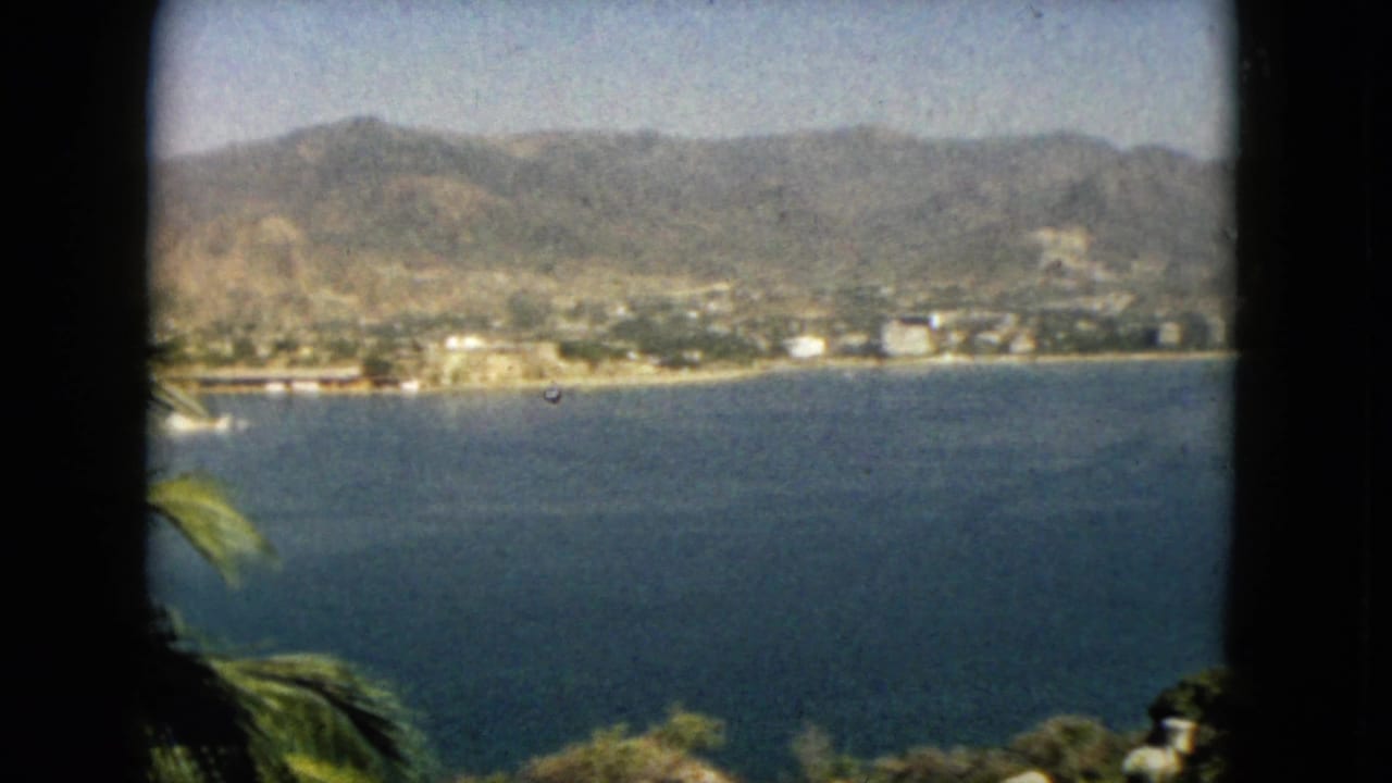 Acapulco, Mexico bay view on 8mm film, with a distant coastal strip, rugged mountains, blue water, and foreground palms.