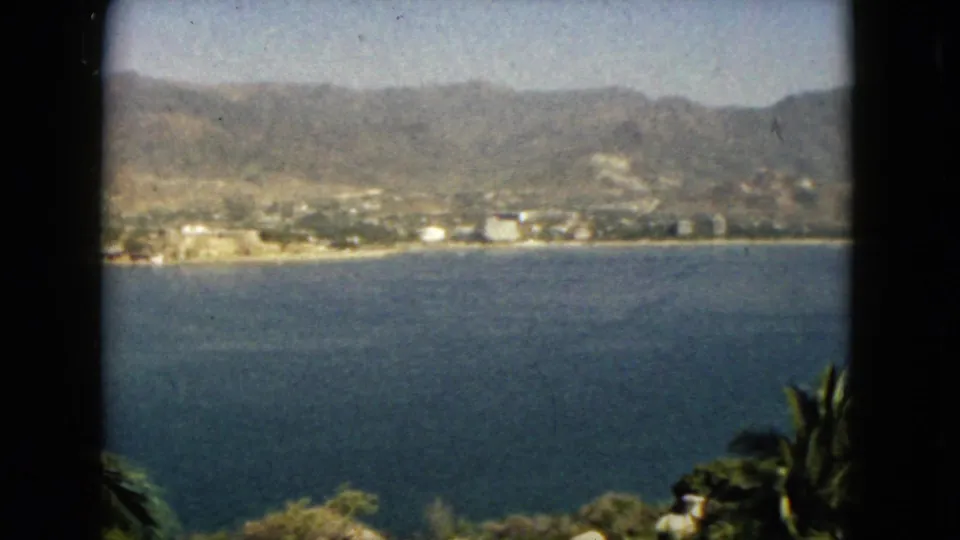Vintage 1962 Acapulco frame looking over calm bay water toward a coastal town and hazy mountain backdrop.