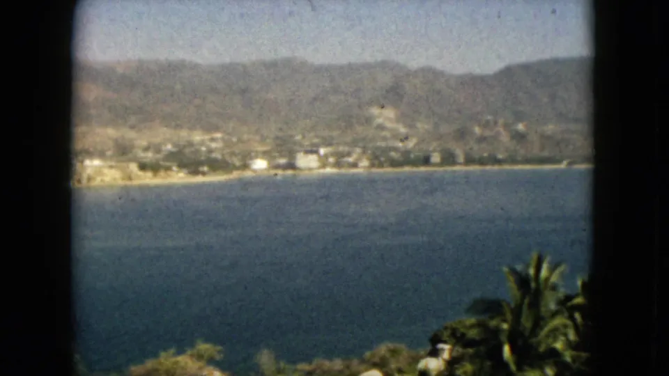 Elevated 8mm home-movie view of Acapulco bay, with mountain ridges, distant waterfront buildings, and palm trees in the foreground.