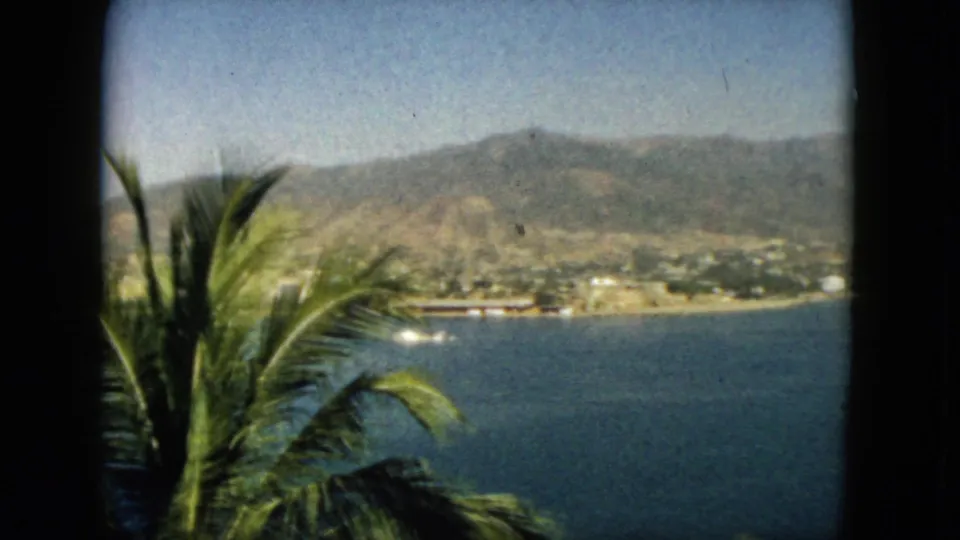 1962 Acapulco home-movie view showing bay water, palms at the edge of frame, shoreline buildings, and mountains.