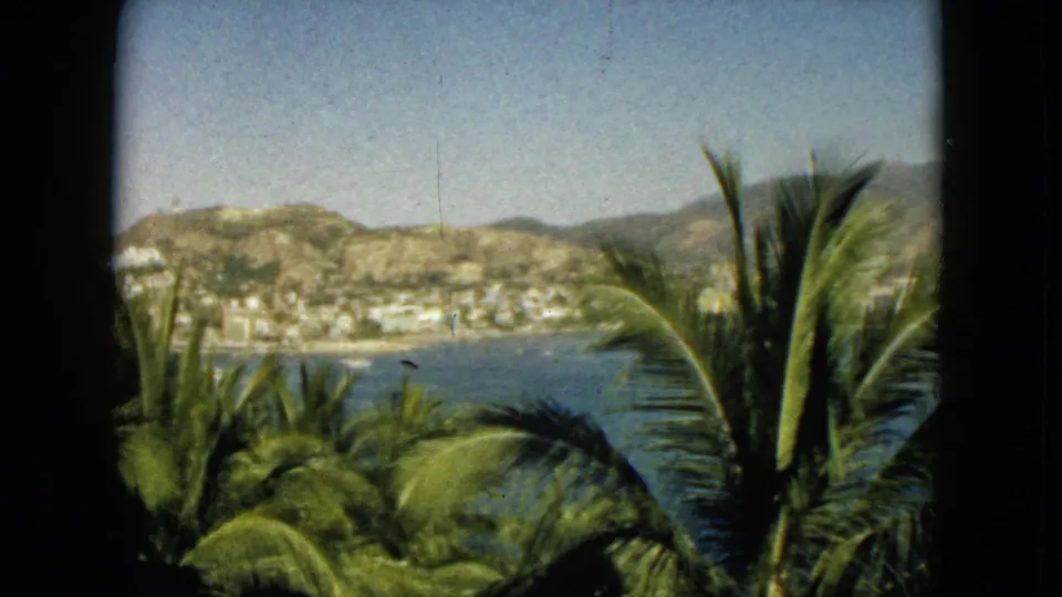 1962 Acapulco 8mm frame showing palms below a window edge, with blue water, clustered shoreline buildings, and hills in the distance.