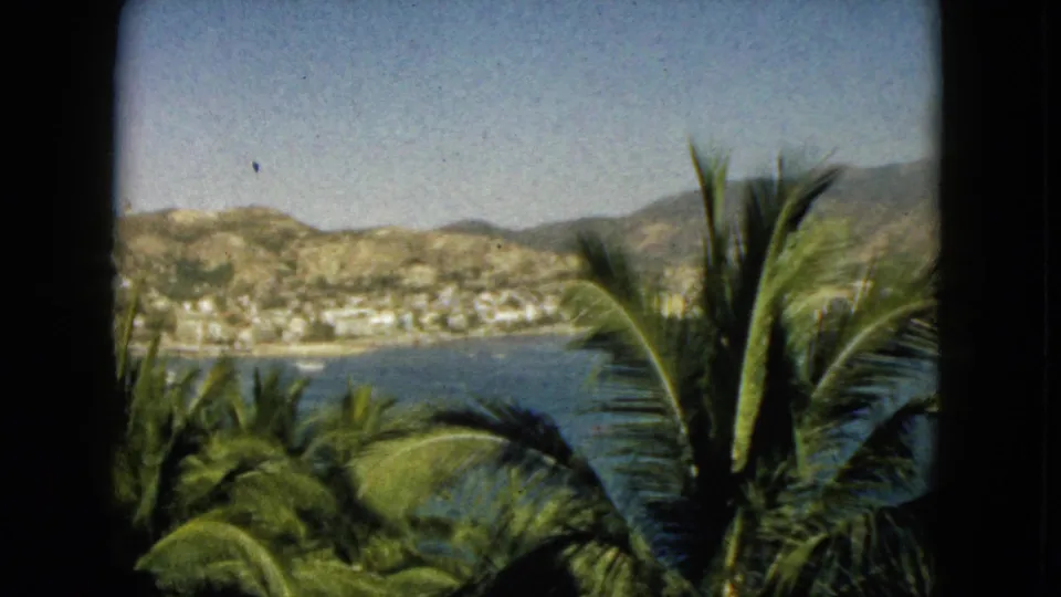 Final 1962 Acapulco frame with a dark window border, palm fronds, bay water, shoreline buildings, distant hills, and open sky.