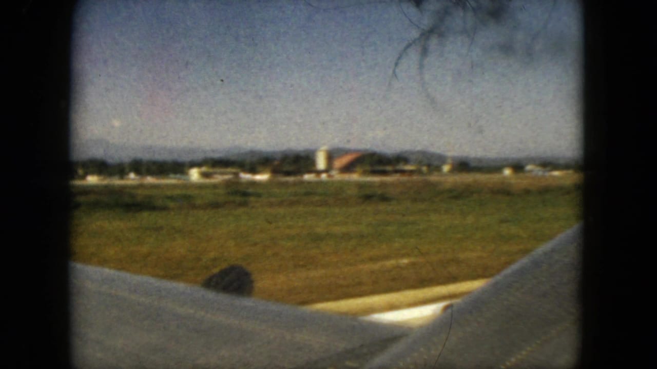From an airplane window in the Acapulco travel footage, the wing crosses the foreground with open ground, low buildings, and hills in the distance.