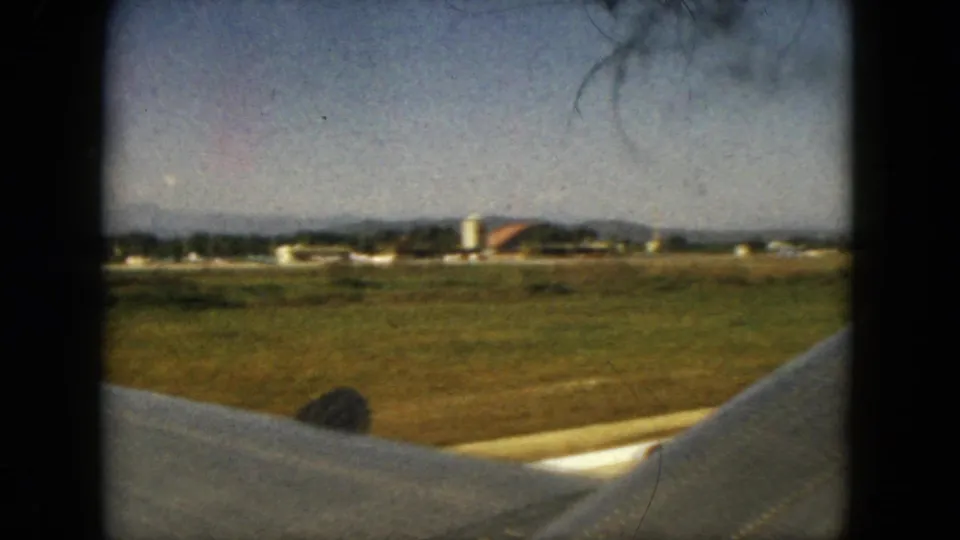 From an airplane window in the Acapulco travel footage, the wing crosses the foreground with open ground, low buildings, and hills in the distance.