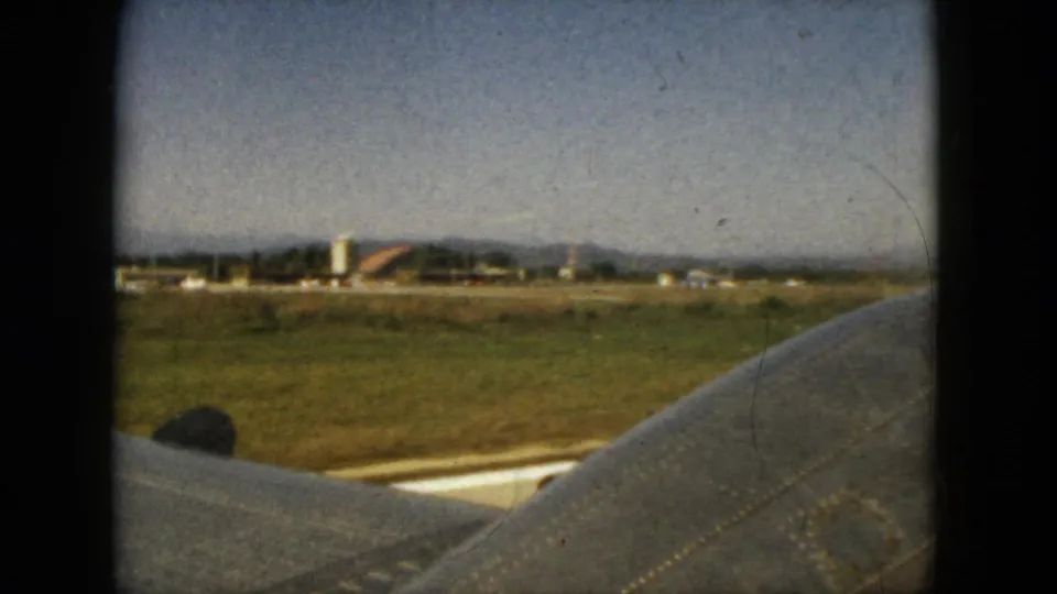 A slightly lower airplane-window angle shows the wing, green-brown field, small distant buildings, and hills under a clear sky.