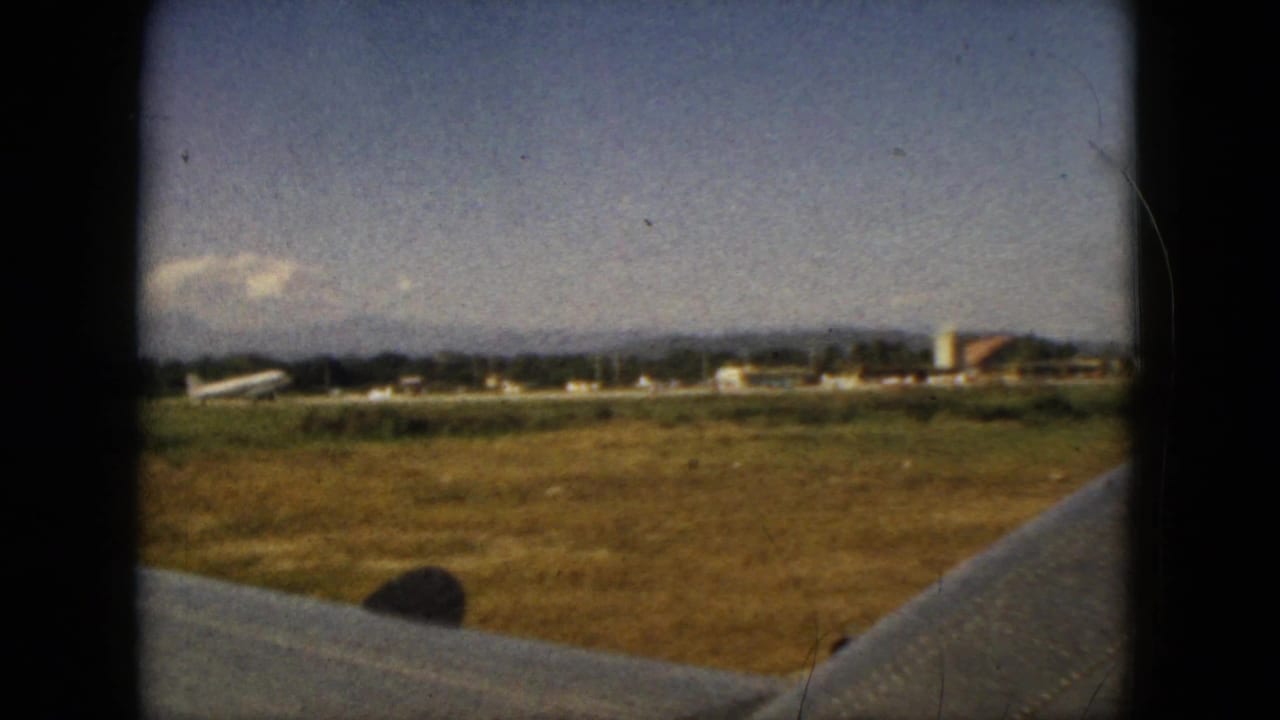 Airplane wing in foreground with grassy runway, distant buildings, mountains, and clouds.