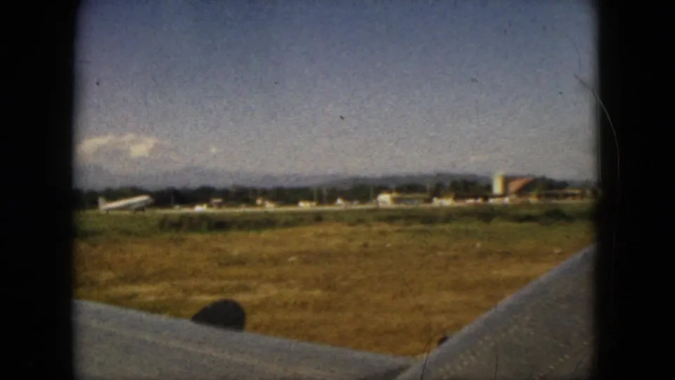 Passenger-window view in Acapulco, Mexico, showing an airplane wing over runway grass with distant airport structures and mountains.