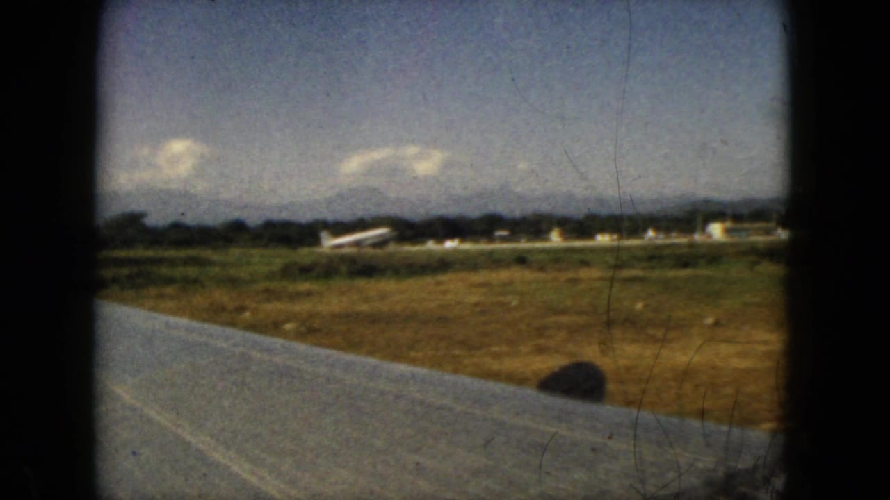 Wing edge over runway grass with a small aircraft and airport buildings in the distance.