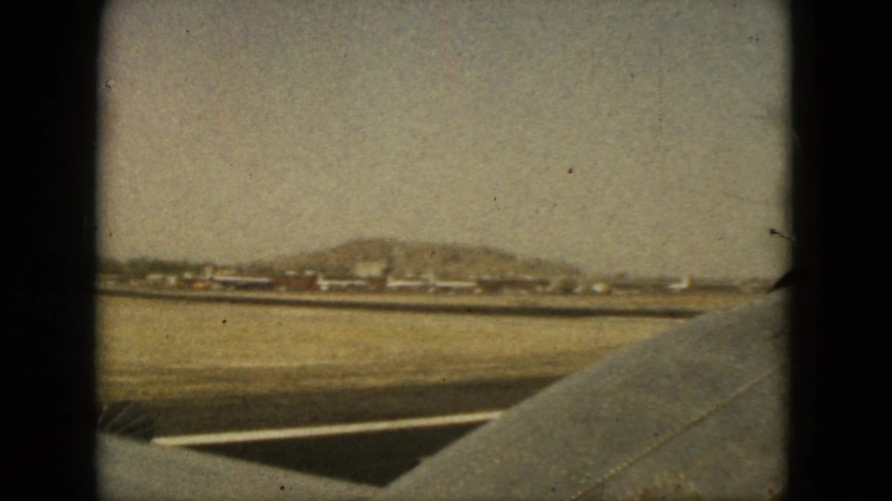 Airplane wing over a runway or ground strip with distant low hills and buildings.