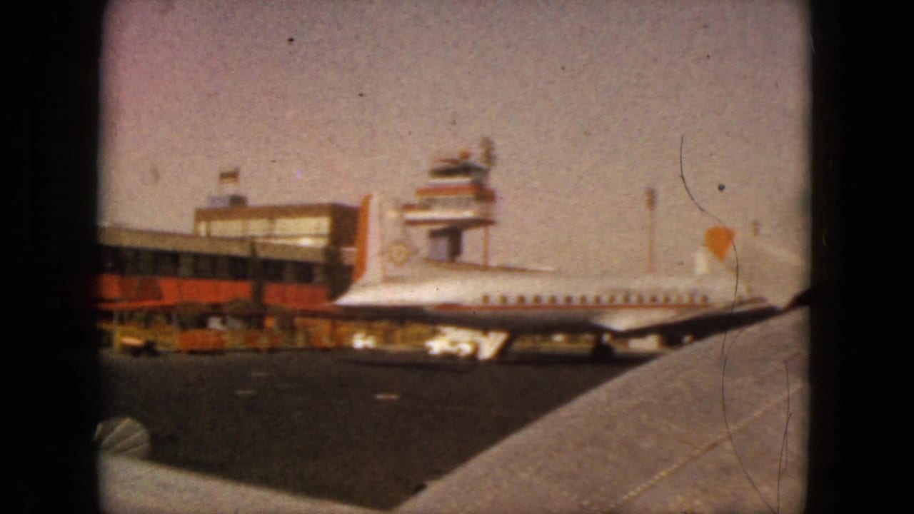 Propeller airliner parked near an airport terminal and control tower, viewed past an airplane wing.